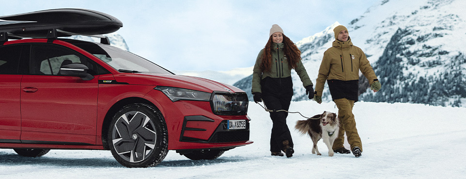 Ein junges Päärchen geht mit Ihrem Hund im Schnee spezieren und Ihr Skoda steht daneben..